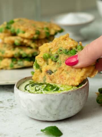 Image of a pea and tofu fritter being dunked in a creamy dipping sauce. There is a stack of fritters behind on a plate.