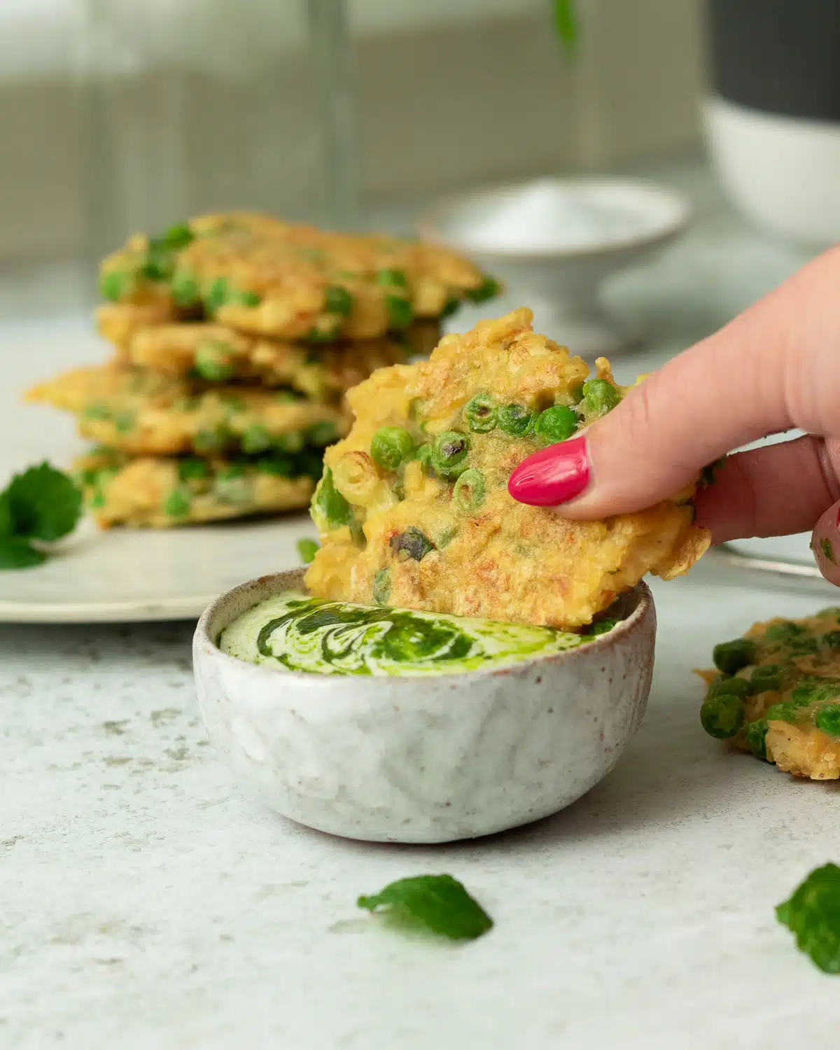 Image of a pea and tofu fritter being dunked in a creamy dipping sauce. There is a stack of fritters behind on a plate. 
