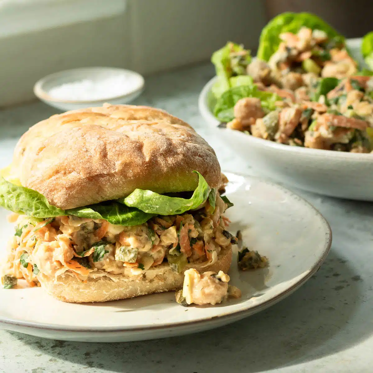 A plate with a bun with chickpea tuna salad on it. There is a plate in the background with lettuce cups filled with chickpea tuna salad too.