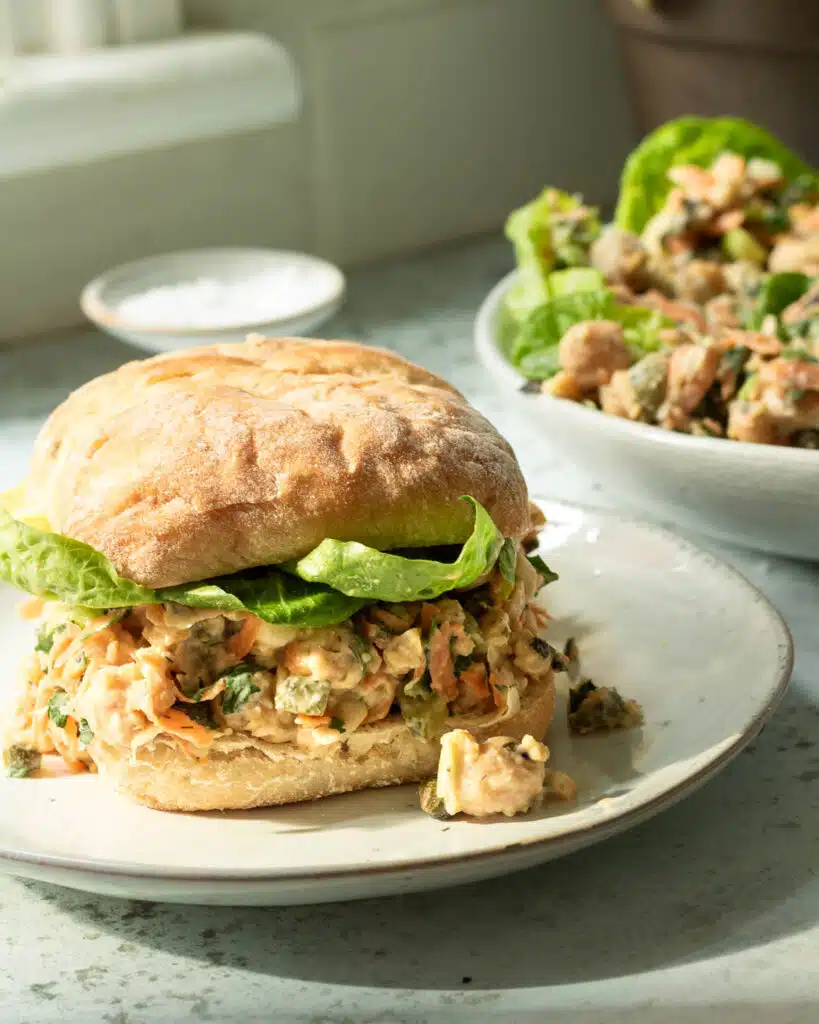 A plate with a bun with chickpea tuna salad on it. There is a plate in the background with lettuce cups filled with chickpea tuna salad too.