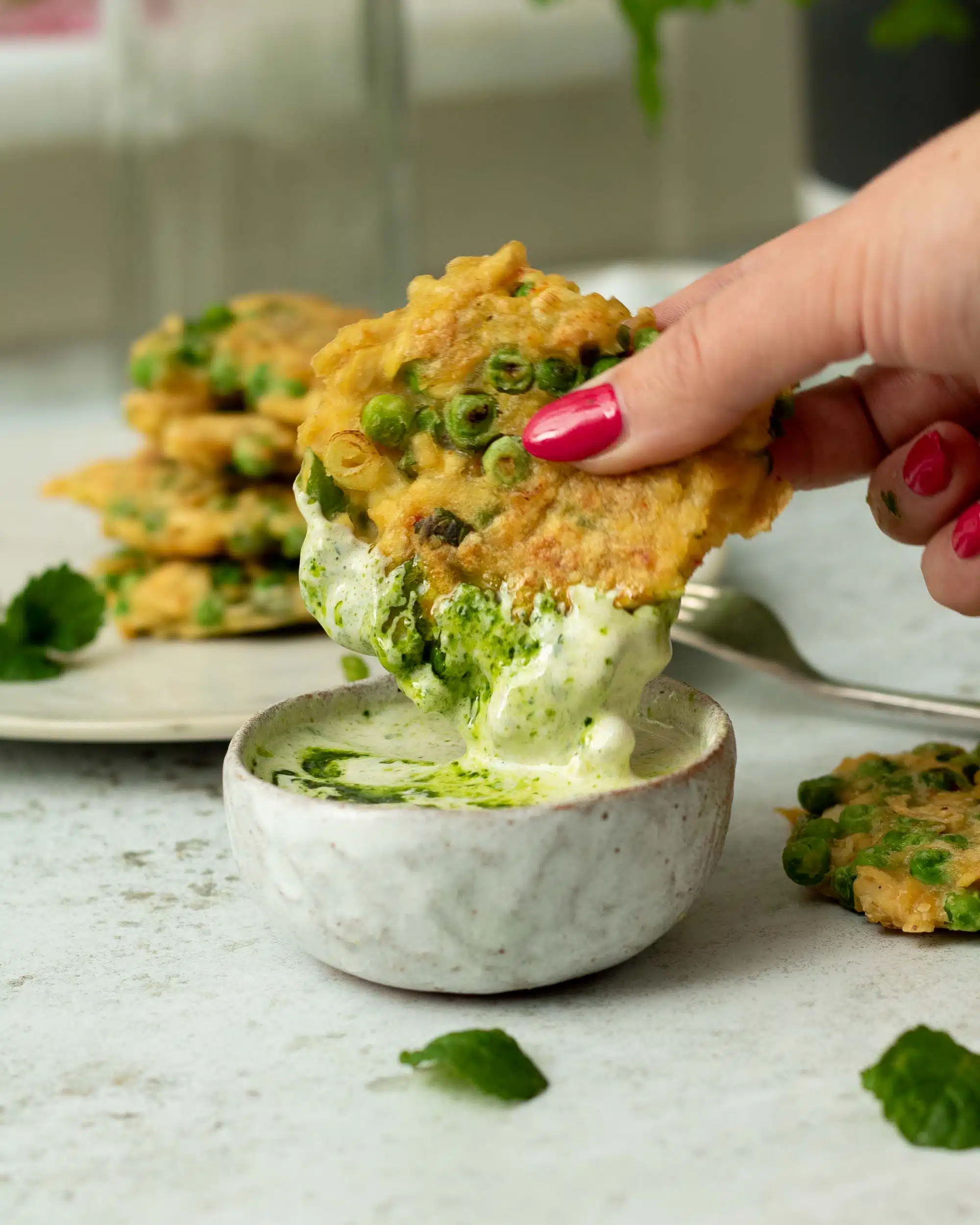 A veggie fritter being dipped into a small bowl of the tahini herb oil. There is another stack of fritters behind.