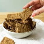 A bowl of lentil flaxseed crackers on a napkin. There is a hand reaching in to pick up a cracker.