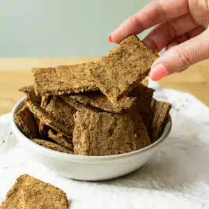 A bowl of lentil flaxseed crackers on a napkin. There is a hand reaching in to pick up a cracker.