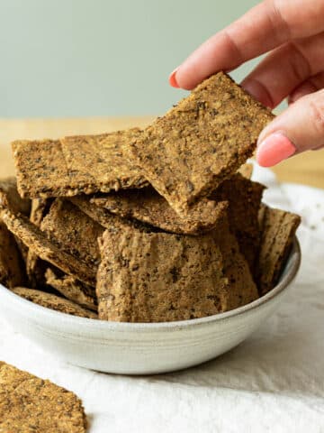 A bowl of lentil flaxseed crackers on a napkin. There is a hand reaching in to pick up a cracker.