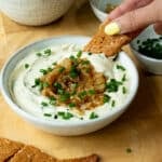 A bowl of creamy onion dip, topped with caramelised onions and chives. There is a hand reaching in and scooping up some dip with a cracker.