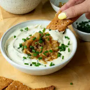 A bowl of creamy onion dip, topped with caramelised onions and chives. There is a hand reaching in and scooping up some dip with a cracker.