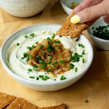 A bowl of creamy onion dip, topped with caramelised onions and chives. There is a hand reaching in and scooping up some dip with a cracker.