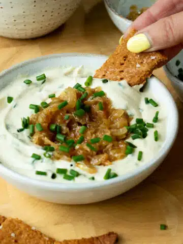 A bowl of creamy onion dip, topped with caramelised onions and chives. There is a hand reaching in and scooping up some dip with a cracker.