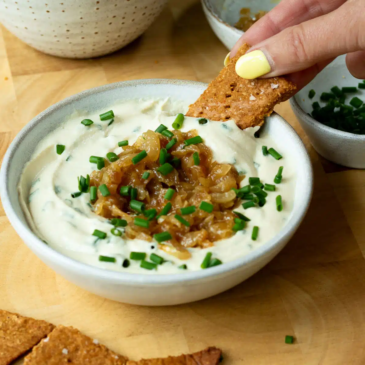 A bowl of creamy onion dip, topped with caramelised onions and chives. There is a hand reaching in and scooping up some dip with a cracker.