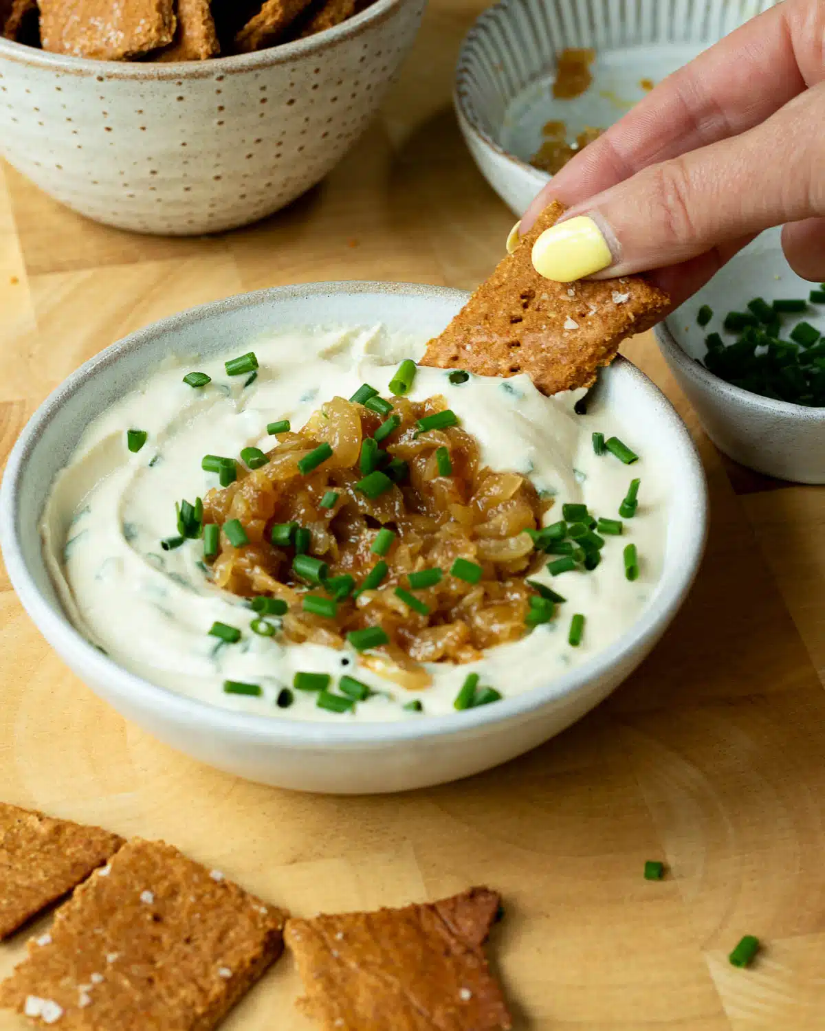 A bowl of creamy onion dip, topped with caramelised onions and chives. There is a hand reaching in and scooping up some dip with a cracker.