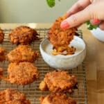 Carrot and Tempeh fritters on a cooling rack with a bowl of tahini dip. There is hand reaching in to dip a fritter into the dip.