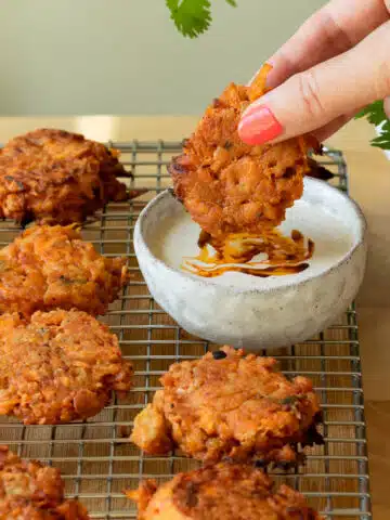 Carrot and Tempeh fritters on a cooling rack with a bowl of tahini dip. There is hand reaching in to dip a fritter into the dip.