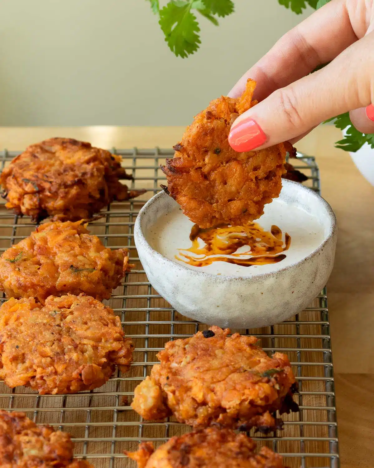 Carrot and Tempeh fritters on a cooling rack with a bowl of tahini dip. There is hand reaching in to dip a fritter into the dip.