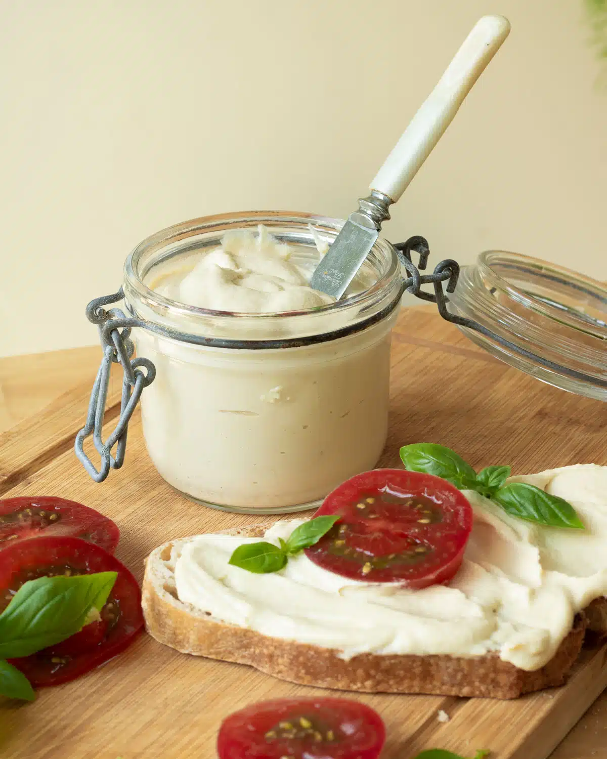 Vegan Cashew cream cheese in a pot with a piece of toast with cream cheese on it witha slice of tomato in front of the pot.
