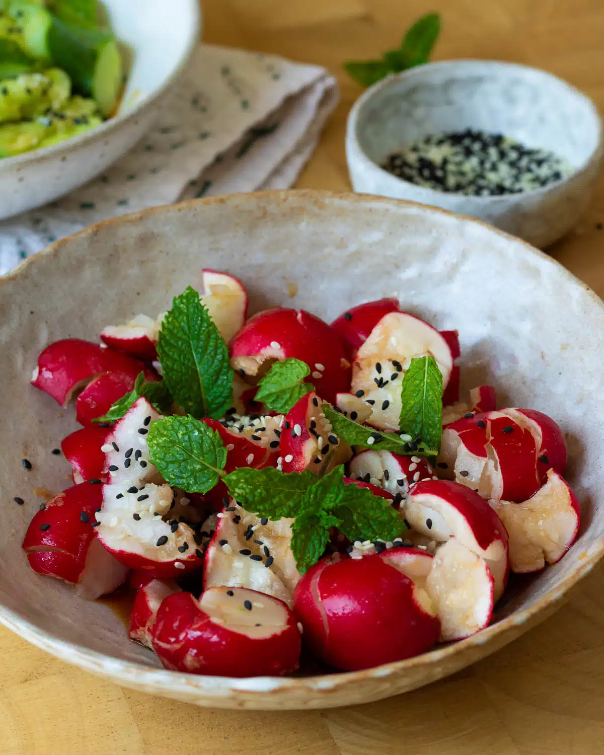 A bowl of smashed radish salad ready to eat. There are mint leaves on top and a bowl of sesame seeds behind.