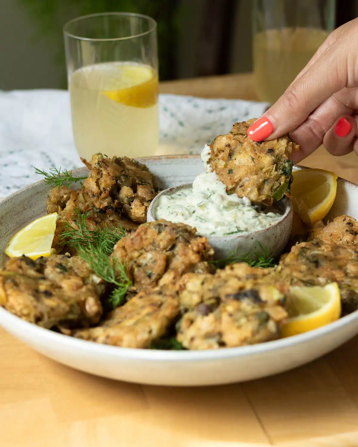 A plate with vegan crab cakes on it, with some tartare sauce. A hand is reaching in and dipping a crab cake in the sauce.