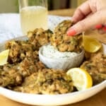 A plate with vegan crab cakes on it, with some tartare sauce. A hand is reaching in and dipping a crab cake in the sauce.