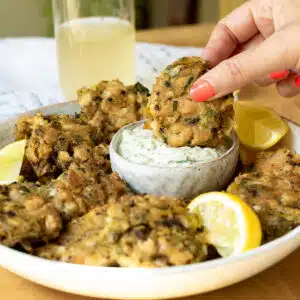 A plate with vegan crab cakes on it, with some tartare sauce. A hand is reaching in and dipping a crab cake in the sauce.