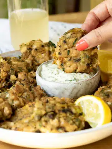 A plate with vegan crab cakes on it, with some tartare sauce. A hand is reaching in and dipping a crab cake in the sauce.