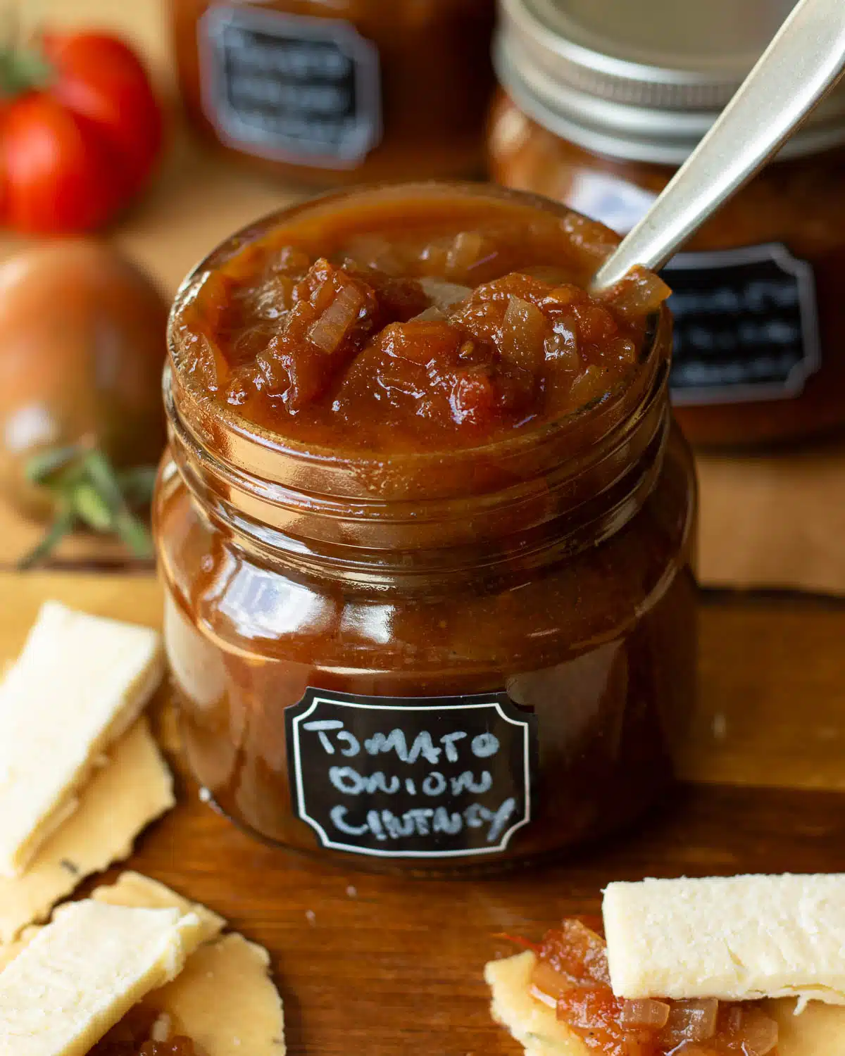 A jar of tomato and onion chutney ready to use, with some cheese and crackers in the background. 