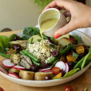 A hand is pouring a vinaigrette salad dressing over a plate of vegan nicoise salad.