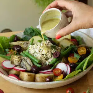 A hand is pouring a vinaigrette salad dressing over a plate of vegan nicoise salad.