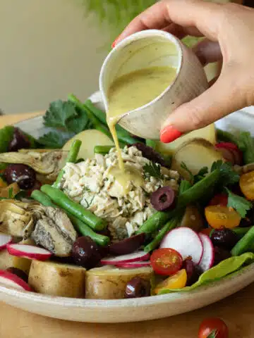 A hand is pouring a vinaigrette salad dressing over a plate of vegan nicoise salad.