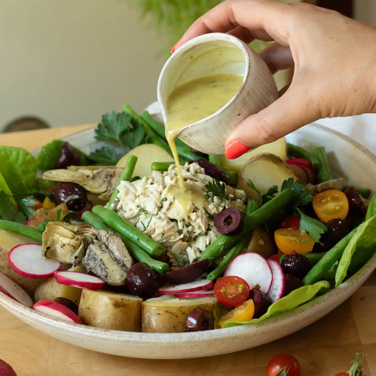 A hand is pouring a vinaigrette salad dressing over a plate of vegan nicoise salad.