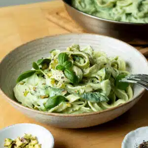 A bowl of lemon basil zucchini pasta ready to eat with a pan of the pasta in the background.