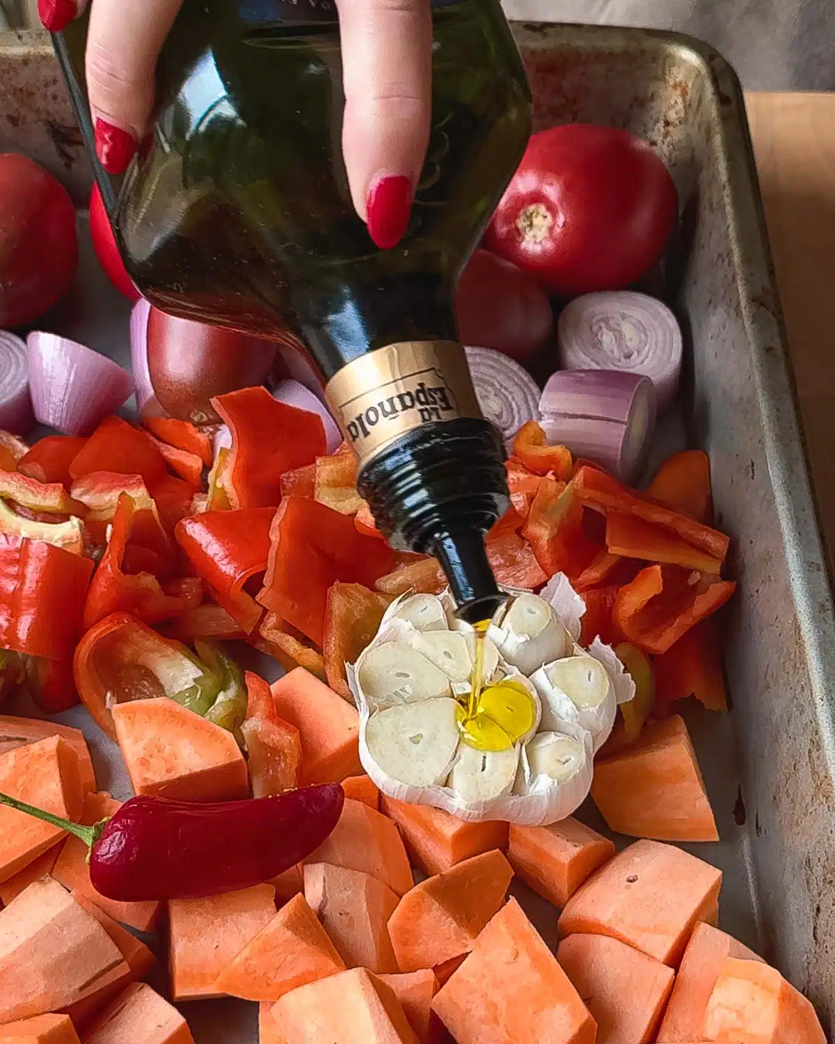 A baking tray with chopped up sweet potatoes, onions, tomatoes and a whole clove of garlic with it's top chopped off, with olive oil being drizzled over it.