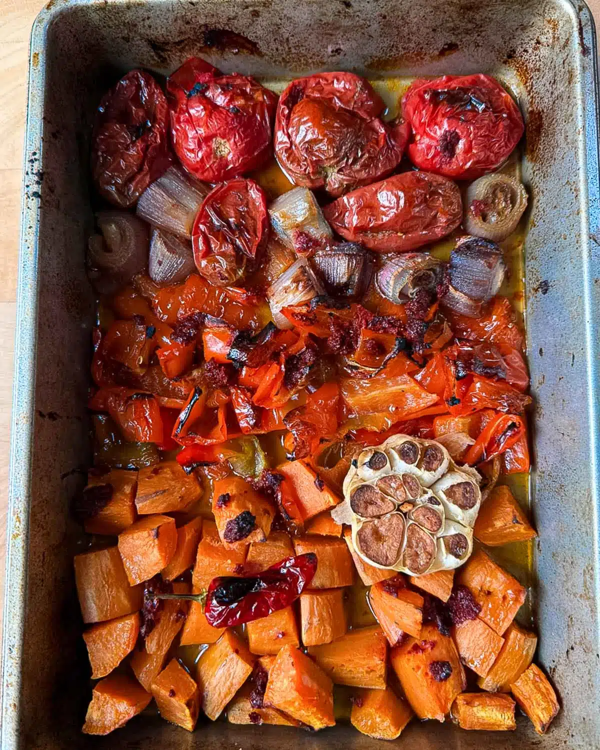 A baking tray with chopped up sweet potatoes, onions, tomatoes and a whole clove of garlic with it's top chopped off, that has been roasted until caramelised and is ready to blend into soup.